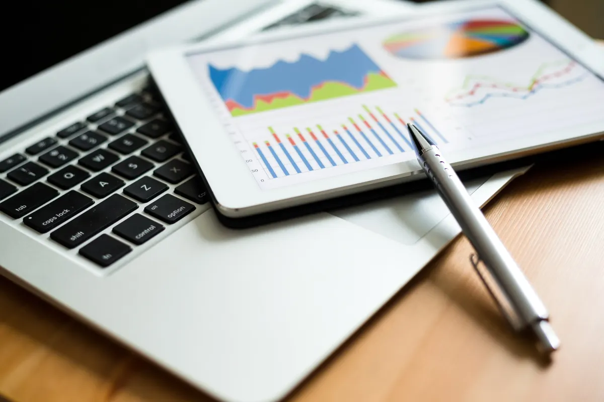 Tablet displaying business analytics charts and graphs alongside a laptop and pen on a wooden desk.