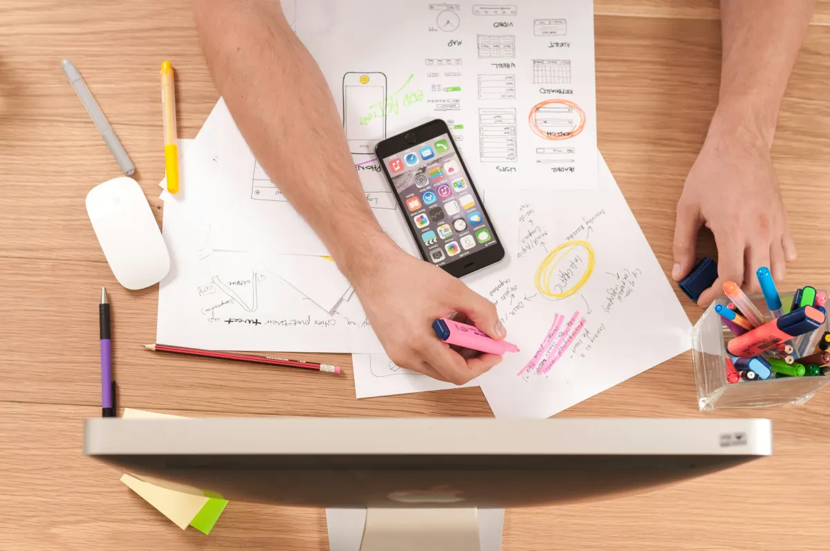 Overhead view of hands working on a desk with design sketches, a smartphone, and creative tools, highlighting content creation.