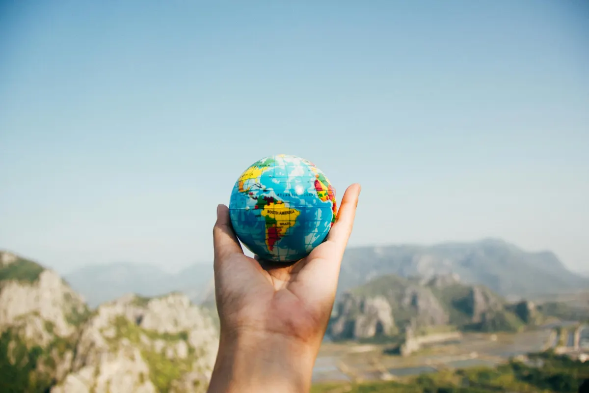 A hand holding a small globe against a backdrop of mountains and a clear sky, symbolizing global reach or environmental care.