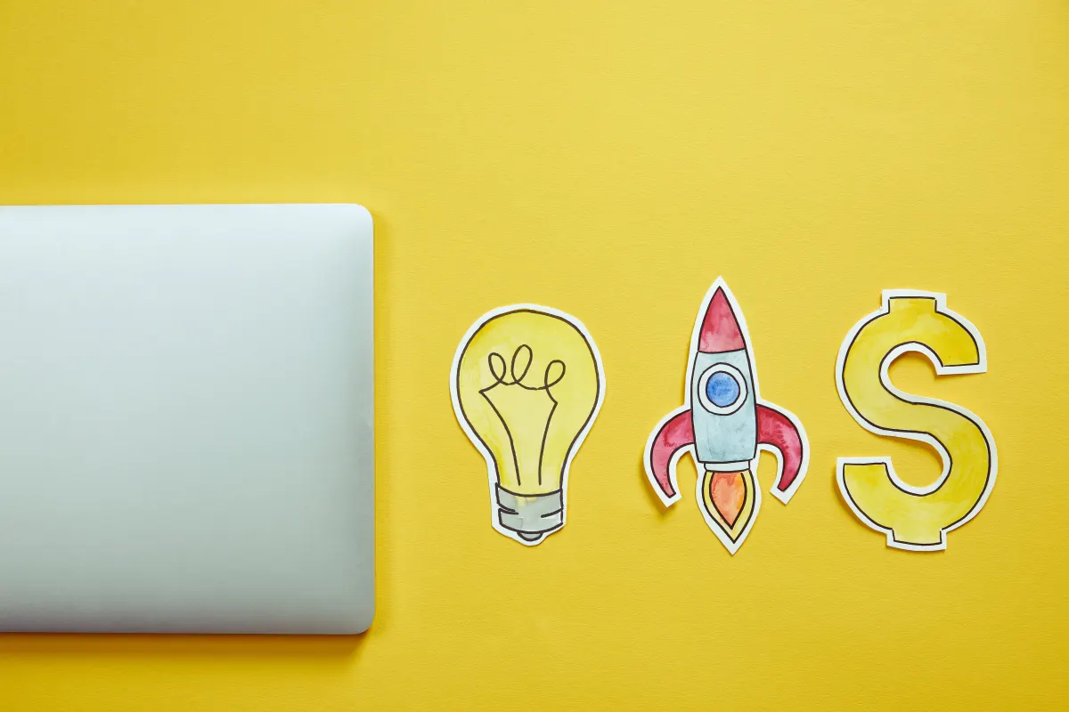 Top view of a laptop and hand-drawn paper cutouts of a lightbulb, a rocket, and a dollar sign on a yellow background, symbolizing innovation, growth, and financial success.
